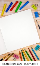 Blank page of open sketchbook with art tools on wooden table. Artistic table top view photo. Drawing or sketch mockup on square notebook page. Blank paper for painting. Crayon and pencil for kids art