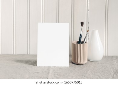 Blank paper card, invitatation mockup. Artistic stationery scene with paint brushes, pencils in ceramic holder and glass vase. Creative table composition, white wooden wall background. - Powered by Shutterstock