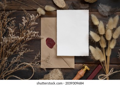 Blank paper card on sealed envelope and wooden table with dried plants, top view. Boho mock-up scene with greeting card template