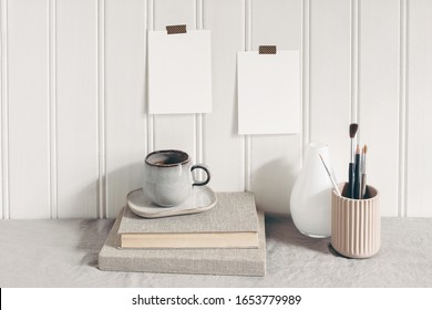 Blank paper cards taped on white wooden wall. Stationery mockup scene with paint brushes, pencils in ceramic holder, cup of coffee and books on linen tablecloth. Artistic scene. Creative background - Powered by Shutterstock