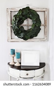 Blank Photo Frame Sitting On A Rustic Half Moon Table Table Below An Old Farmhouse Window Decorated With A Homemade Magnolia Leaf Wreath On An Interior Wall.