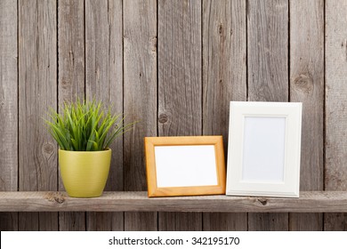 Blank Photo Frames And Plant On Shelf In Front Of Wooden Wall