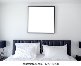 Blank photo square frame mockup on white wall above the bed with white clean pillows in the bedroom.