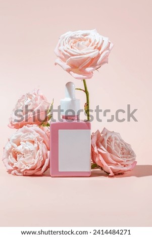 Blank pink glass essential oil bottle with pipette on pink background decorated blooming rose flowers. Skin care concept with natural cosmetics.