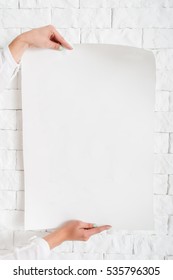 Blank Poster In Female Hands Against Wall Mockup. Close-up Of Woman Holding Empty Sheet Of Paper With Free Space For Text, Advertisement, Promo, Warning. Backdrop For Message