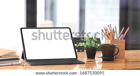 Blank screen computer tablet with keyboard case putting on wooden working desk with pencil holder, coffee cup, potted plant, stack of books and pen over neat living room as background.