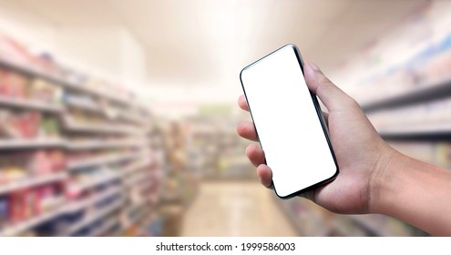 Blank Screen Of Mobile Phone With Hand Holding Phone On Soft Blurry Supermarket Store Background. Of Free Space For Copy And Branding.