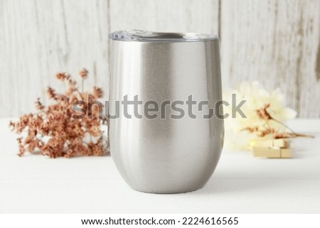 Blank silver wine tumbler with lid mockup in white wood background with red, beige flower. Svg product mockup, svg product display, mockup for svg product, drinkware mockup. Front view, copy space.