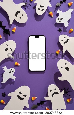 A blank smartphone screen surrounded by paper ghosts, candy corn, bats and spiders for a playful Happy Halloween composition with purple background and mockup space for seasonal designs