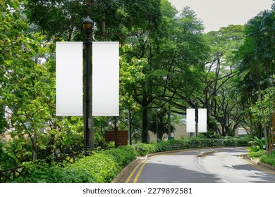 Blank vertical advertising banners on street lampposts; double hanging posters by the road, against lush green trees and plants. For OOH out of home template mock up.
