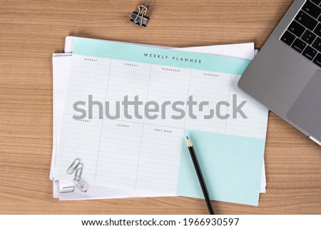 Blank weekly planner notice block on a desk in an office. Empty schedule and a pencil. To-do list and paperclips on the table. Laptop lying down.