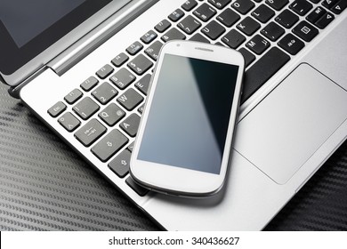 Blank White Business Smartphone With Reflection Lying On A Notebook Keyboard, All Above A Carbon Layer