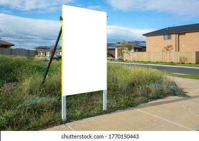 Blank White Empty Mockup Template Of A Real Estate Sign At Front Of A Lot Of Vacant Land In A Suburban Neighbourhood With Some Residential Houses At The Background.