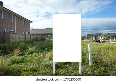 Blank White Empty Mockup Template Of A Real Estate Sign At Front Of A Lot Of Vacant Land On A Suburban Neighbourhood Street With Some Residential Houses At The Background.