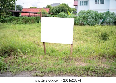 Blank White Empty Mockup Template Of A Real Estate Sign At Front Of A Lot Of Vacant Land In A Suburban Neighbourhood With Some Residential Houses At The Background.