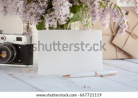 Blank white greeting card with lilac flowers bouquet and envelope with vintage camera on white wooden background. mock up