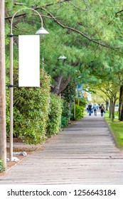 Blank White Isolated Vertical Banner Flag Mockup Signage Sign On A Streetlight. Public City Park Walkway. Unrecognizable People Walking In Far Distance Background.