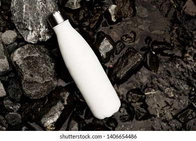A blank white metal water bottle lying on wet black stones.