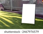 Blank white mockup background texture of a standing easel at the entrance of an entertainment ride in a park. Empty sign board placed outdoor on artificial turf against metal fence.