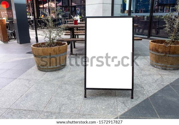 Blank white outdoor advertising stand/sandwich\
board mock up template. Clear street signage board placed by an\
outdoor dinning area of a restaurant. Background texture of standee\
on street.