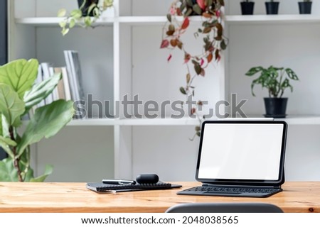 Blank white screen tablet with magic keyboard, gadget and houseon wooden table in living room.