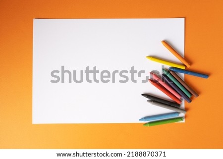 A blank white sheet of paper with wax crayons lies on a orange background.