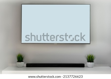 Blank white smart TV screen in grey, bright mockup, front view. Empty telly LED display in living room mock up. Clear panel monitor including sound bar and artificial grass in the pot. 