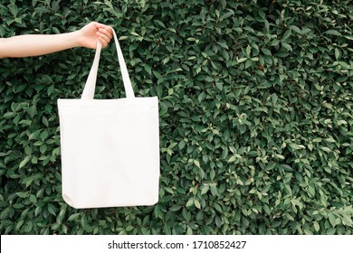 Blank White Tote Bag Canvas Fabric With Handle Mock Up Design. Close Up Of Woman Hand Holding Eco Or Reusable Shopping Bag On Green Leaves Background. No Plastic Bag And Ecology Concept. Copy Space.