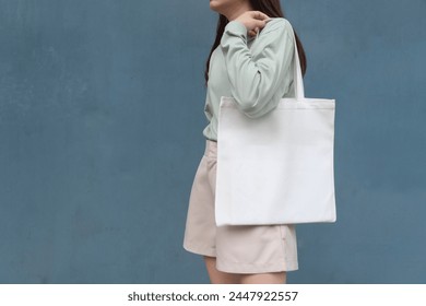 Blank white tote bag canvas fabric with handle mock up design. Close up of woman holding eco or reusable shopping bag against blue metal wall. No plastic bag and ecology concept.