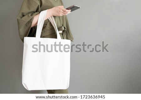 Blank white tote bag canvas fabric with handle mock up design. Close up of woman holding eco or reusable shopping bag and using smartphone near grey wall. No plastic bag and ecology concept.
