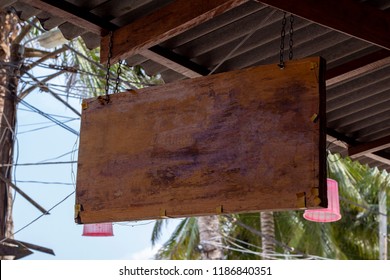 Blank Wooden Board Sign Hang Under Roof. Tropical Island Cafe Or Hotel Sign Mockup. Wooden Board With Palm Leaf Background. Exotic Place For Vacation. Empty Billboard Outdoor. Rustic Board Texture