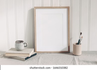 Blank wooden frame mockup with paint brushes, pencils in ceramic holder, cup of coffee and books on linen tablecloth. Artistic scene. Creative table background.. Poster product design, wooden wall. - Powered by Shutterstock