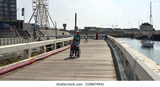 Blankenberge, West-Flanders Belgium - September 04, 2022: Older Couple, Woman In A Wheel Chair Moving On The Pier. Other Tourists Walking Or Looking To The Sailing Boats On The Pier