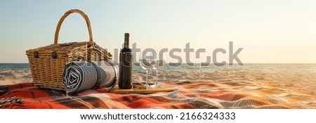 Blanket with picnic basket, bottle of wine and glasses on sandy beach near sea, space for text. Banner design