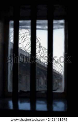 A bleak and dismal view through a prison window. The bars over the window with the only view of coils of barbed wire on a wall is symbolic of the lack of freedom for prisoners.