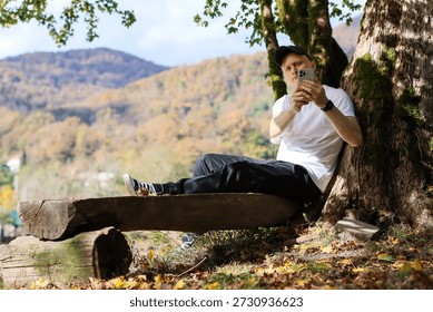 Blending tech with tranquility, man in casual wear takes photo on phone while lounging in autumn forest under dappled sunlight