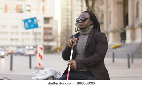Blind black man sitting on the bench and holding walking long cane. High quality photo