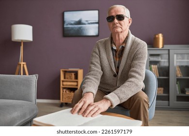 Blind senior man in eyeglasses reading a book in Braille at table in the living room - Powered by Shutterstock