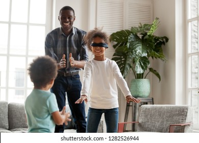 Blindfolded Little Girl Playing Hide And Seek At Home With African Dad And Kid Brother, Happy Black Dad And 2 Mixed Race Children Laughing Enjoy Active Game Together, Family Having Fun In Living Room