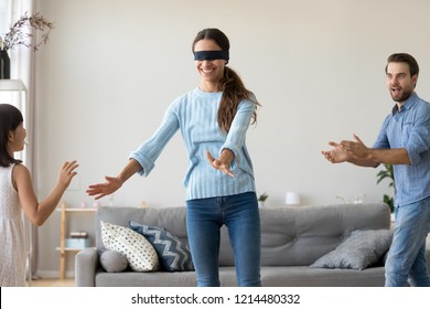 Blindfolded mother catching little daughter and husband playing hide and seek in living room at home, cheerful kid having fun with mommy and daddy. Weekend leisure activities and family games concept