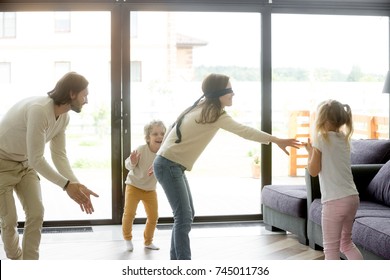 Blindfolded mother playing hide and seek game with father and kids hiding clapping hands at home, happy family having fun together on leisure in living room, parents children activities on weekend