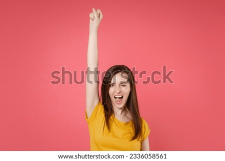 Blinking cheerful funny beautiful young brunette woman 20s wearing yellow casual t-shirt posing standing rising hand up clenching fist like winner isolated on pink color background studio portrait