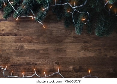 Blinking Christmas Decorative Light String With Green Pine Leaves On Wood Table Background, Top View Border Design With Copy Space