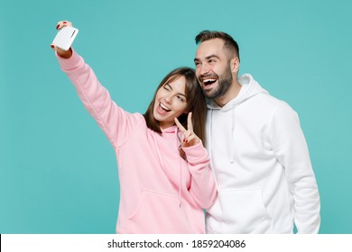 Blinking funny young couple friends man woman in white pink casual hoodie doing selfie shot on mobile phone showing victory sign hugging isolated on blue turquoise colour background studio portrait