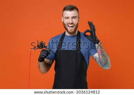 Blinking professional tattooer master artist tattooed man in blue t-shirt apron hold machine black ink in jar, equipment for making tattoo art on body showing OK gesture isolated on brown background