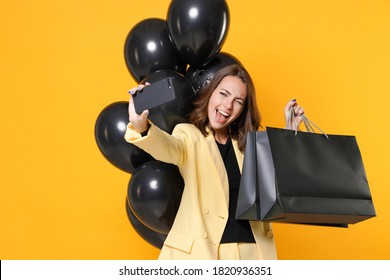 Blinking young woman 20s in suit jacket hold package bags with purchases after shopping bunch of air balloons doing selfie shot on mobile phone isolated on yellow background studio. Black friday sale Foto Stock