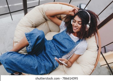 Blissful black young woman in vintage denim clothes chilling on terrace in white headphones. Attractive girl enjoying fresh air on balcony and listening music in earphones in morning.