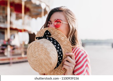 Blissful blonde girl covering face with straw hat while posing in summer day. Outdoor photo of happy young woman in pink sunglasses resting in amusement park.