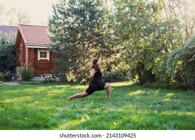 Blissful fat woman doing exercises for stretching legs green grass on backyard of cottage with wooden house and trees in background. Body positivity. Equality. 