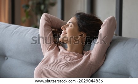 Blissful female closed eyes relaxing alone on comfy sofa at home, close up. Pretty young Latin woman put hands behind head breathing fresh conditioned air inside of modern house, stress-free concept